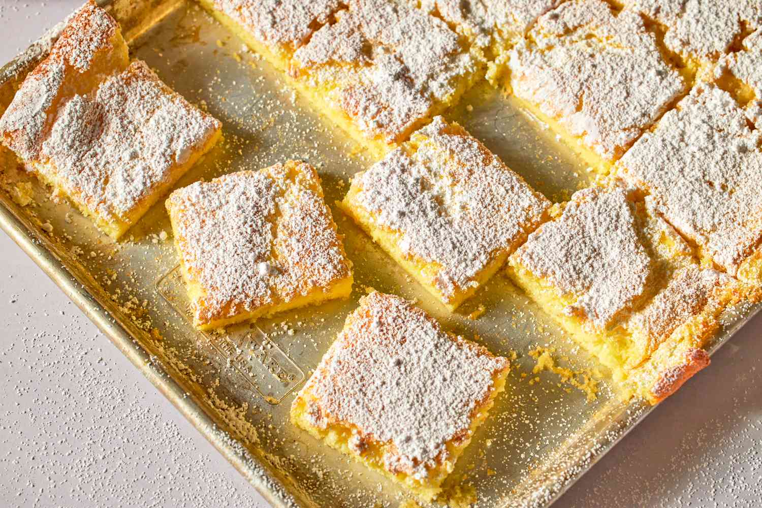 A tray of baked dessert bars dusted with powdered sugar, cut into square servings, displayed on a metallic surface