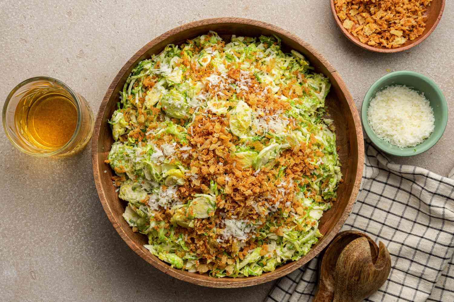 Bowl of Brussels Sprouts Caesar Salad with Lemony Breadcrumbs Next to Serving Utensils on a Table Napkin, a Bowl with More Toppings, a Bowl with Shredded Parmesan, and a Glass
