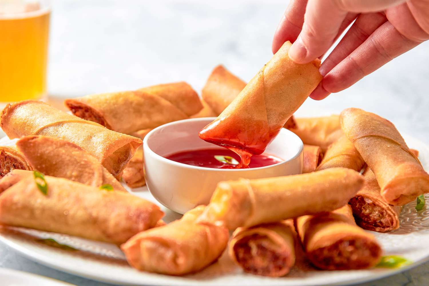 Piece of Lumpia Dipped in a Bowl With Sweet and Sour Sauce Sitting on a Plate With More Lumpia, and in the Background, a Glass With a Fizzy Drink