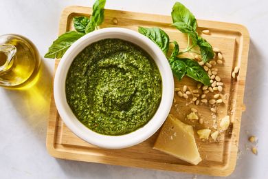 Bowl of pesto on a wooden cutting board with basil leaves pine nuts Parmesan cheese and olive oil nearby