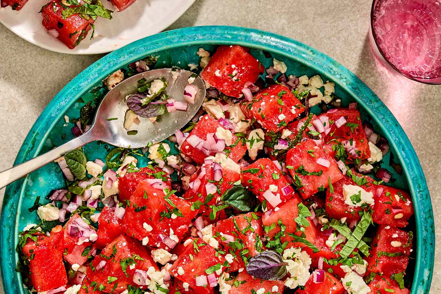 A bowl of watermelon feta salad garnished with herbs and onions, with a spoon resting on the side of the dish