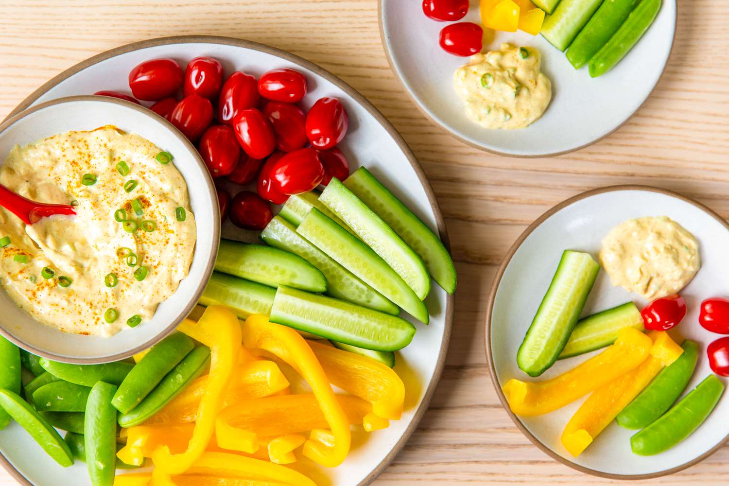A plate of fresh vegetables with a creamy dip, consisting of cherry tomatoes, cucumber sticks, bell pepper strips, and peas