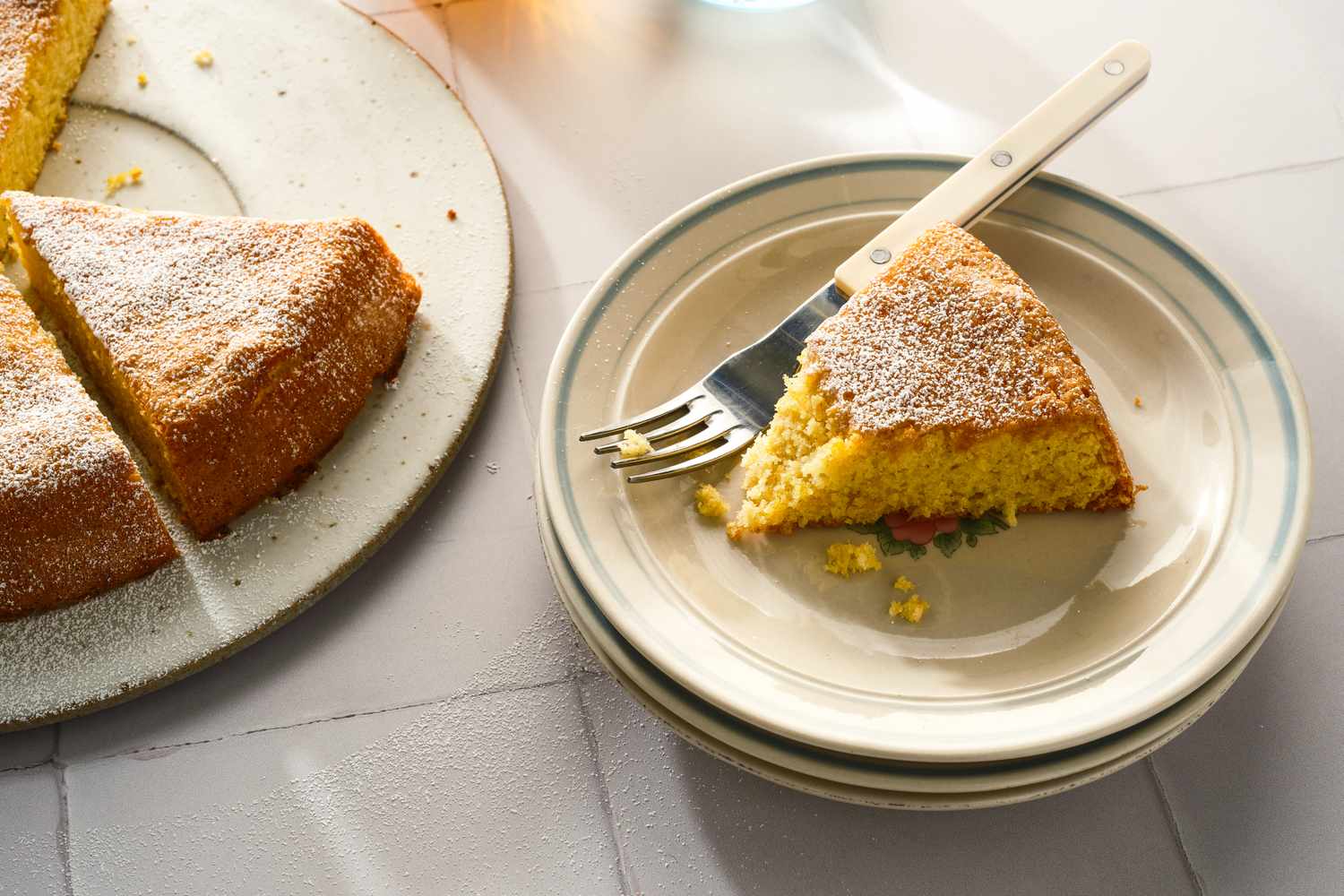 A slice of almond flour lemon cake on a small plate with a fork beside the remaining cake