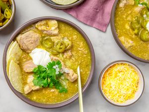 A bowl of pork chile verde garnished with sour cream cilantro and jalapenos accompanied by lime wedges and shredded cheese in bowls