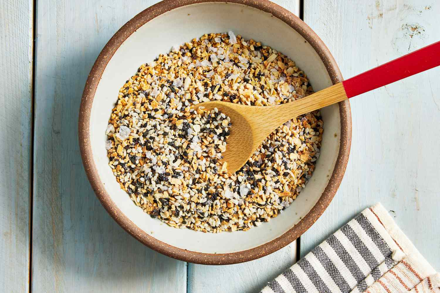 Bowl of Everything Bagel Seasoning With a Wooden Spoon and Next to a Black and White Stripped Kitchen Towel