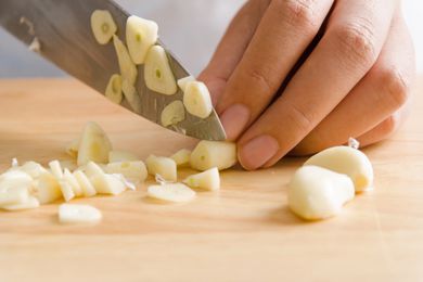 A hand holding a garlic clove with a knife chopping it on a cutting board