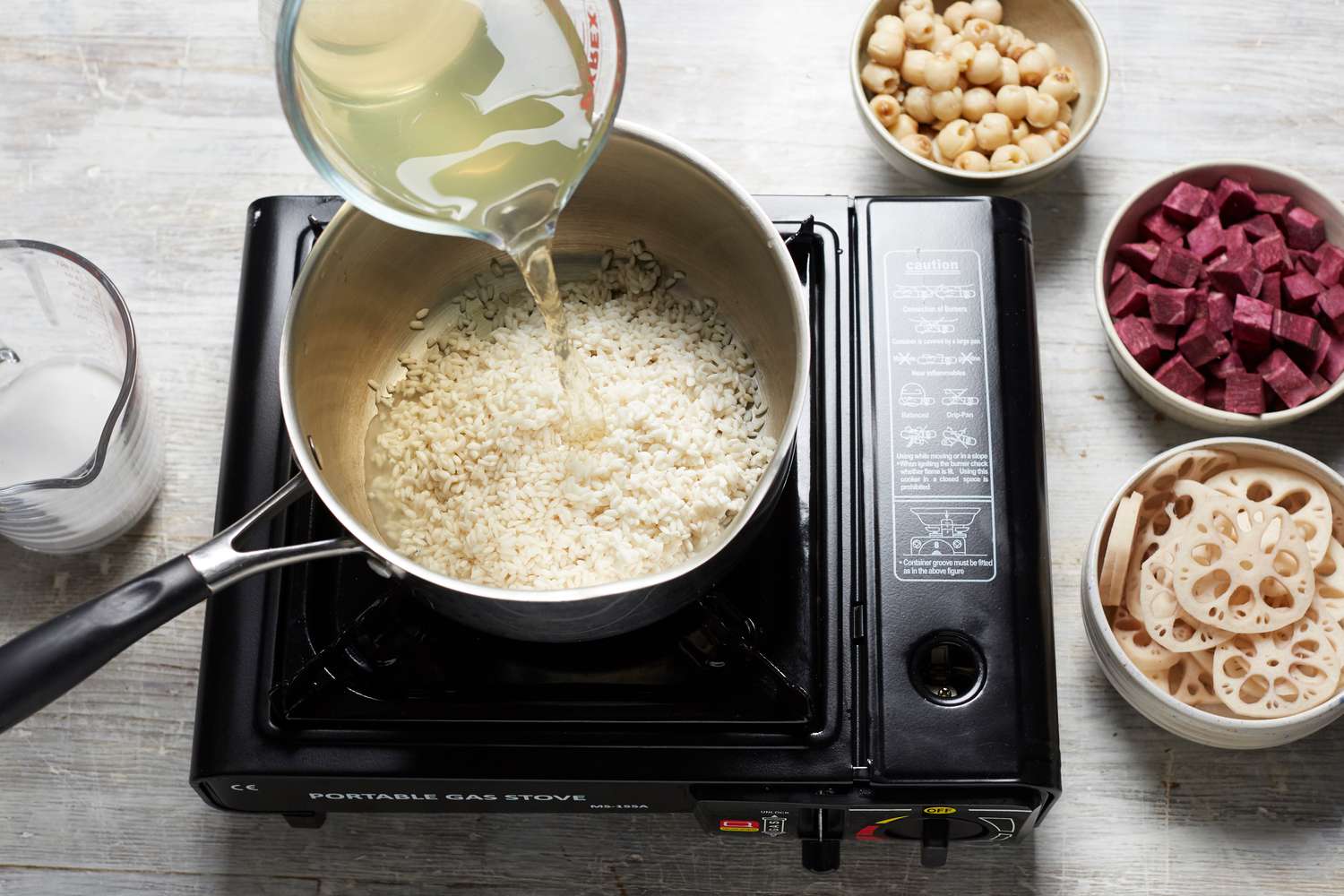 Adding liquid to a pot of rice to soak with bowls of ingredients next to it to make lotus seed, lotus root, and sweet potato rice pudding