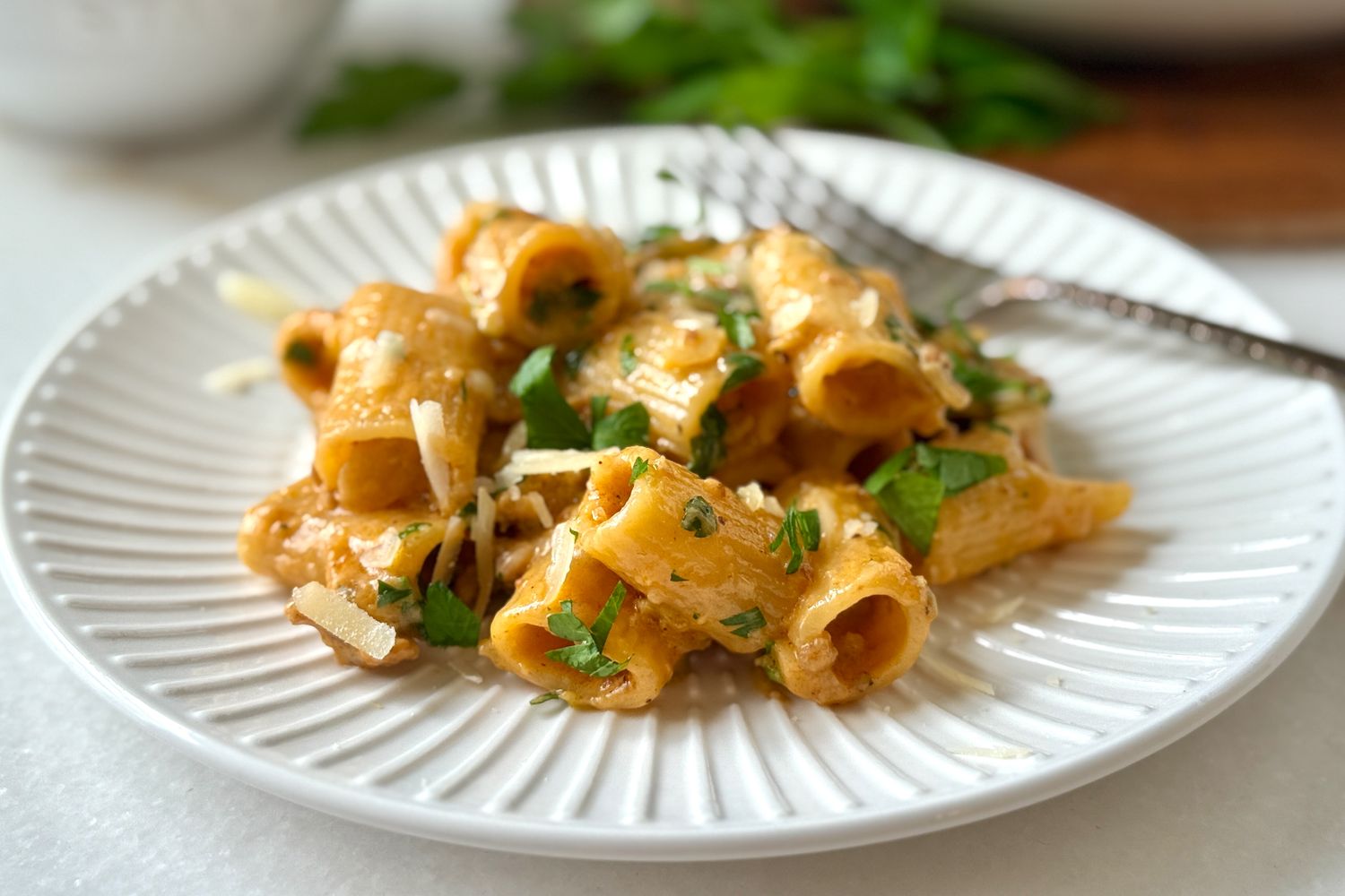 A plate of rigatoni pasta garnished with herbs and grated cheese set on a white fluted dish with a fork