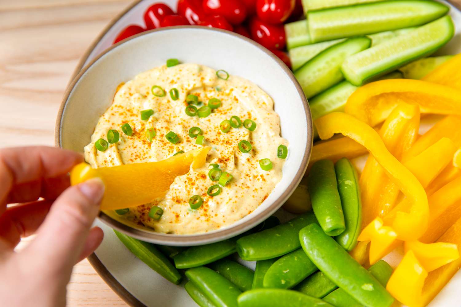 Bowl of curry dip surrounded by vegetables, with a hand dipping a yellow bell pepper slice