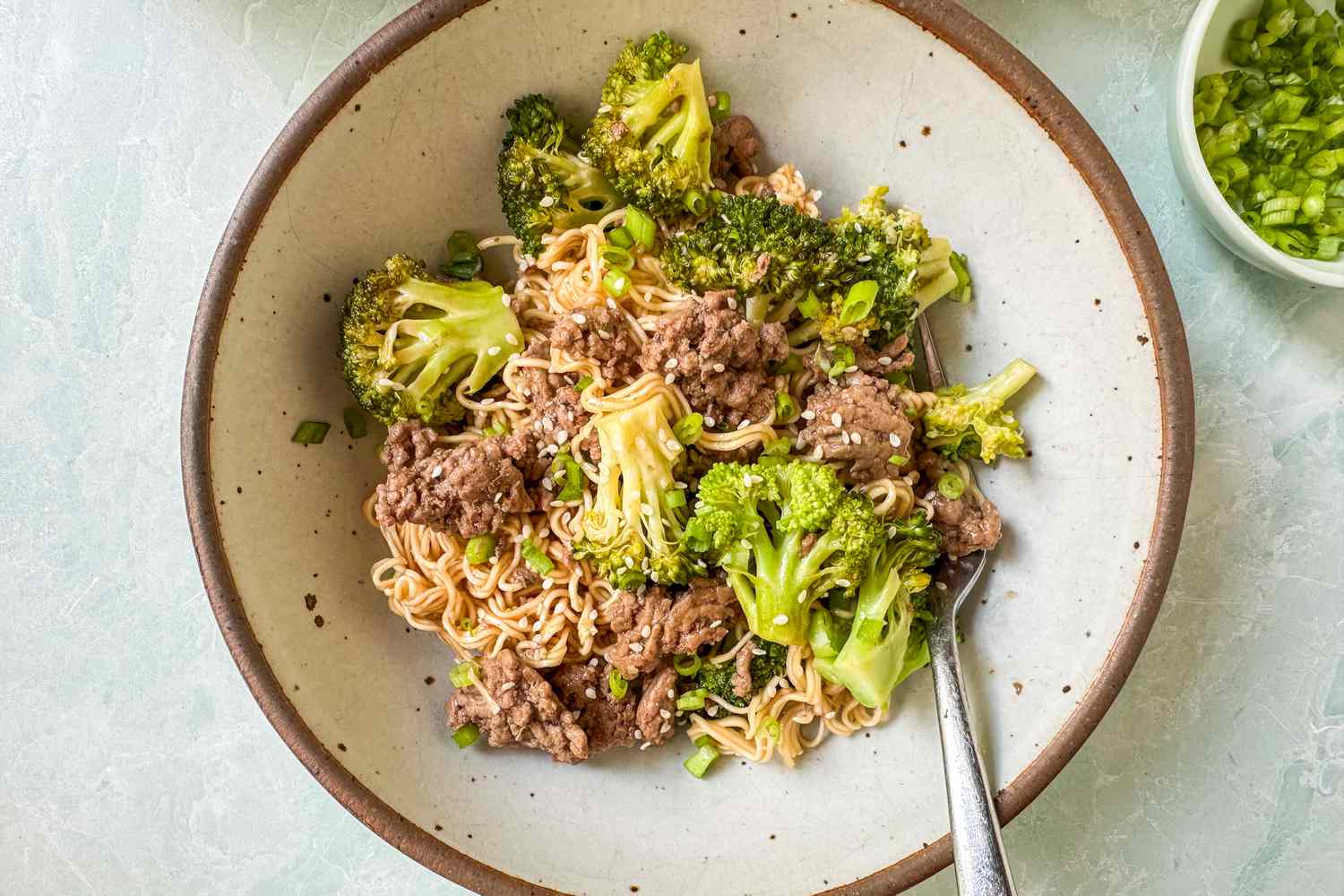 Overhead view of a brown rustic style bowl of ground beef, broccoli, noodles and a fork next to a small bowl of chopped green onions on a light blue background