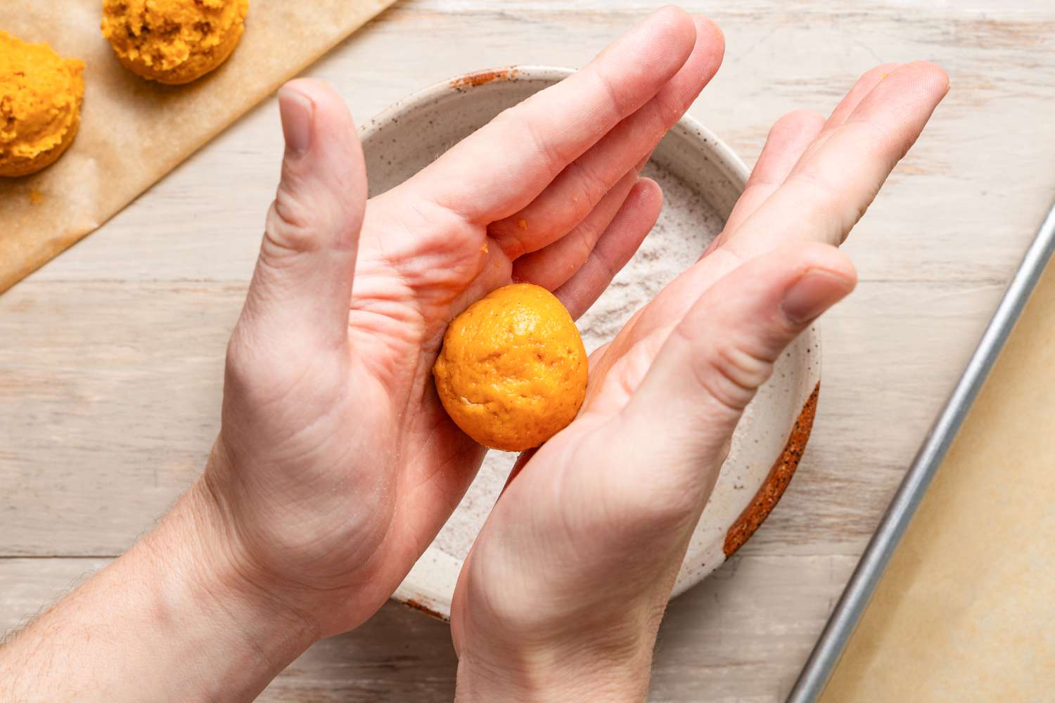 Hands rolling and shaping pumpkin spice cookie dough into a small cookie dough ball, and in the background, more cookie dough balls on a piece of parchment paper, a bowl with pumpkin spice sugar mixture, and a parchment paper lined baking sheet