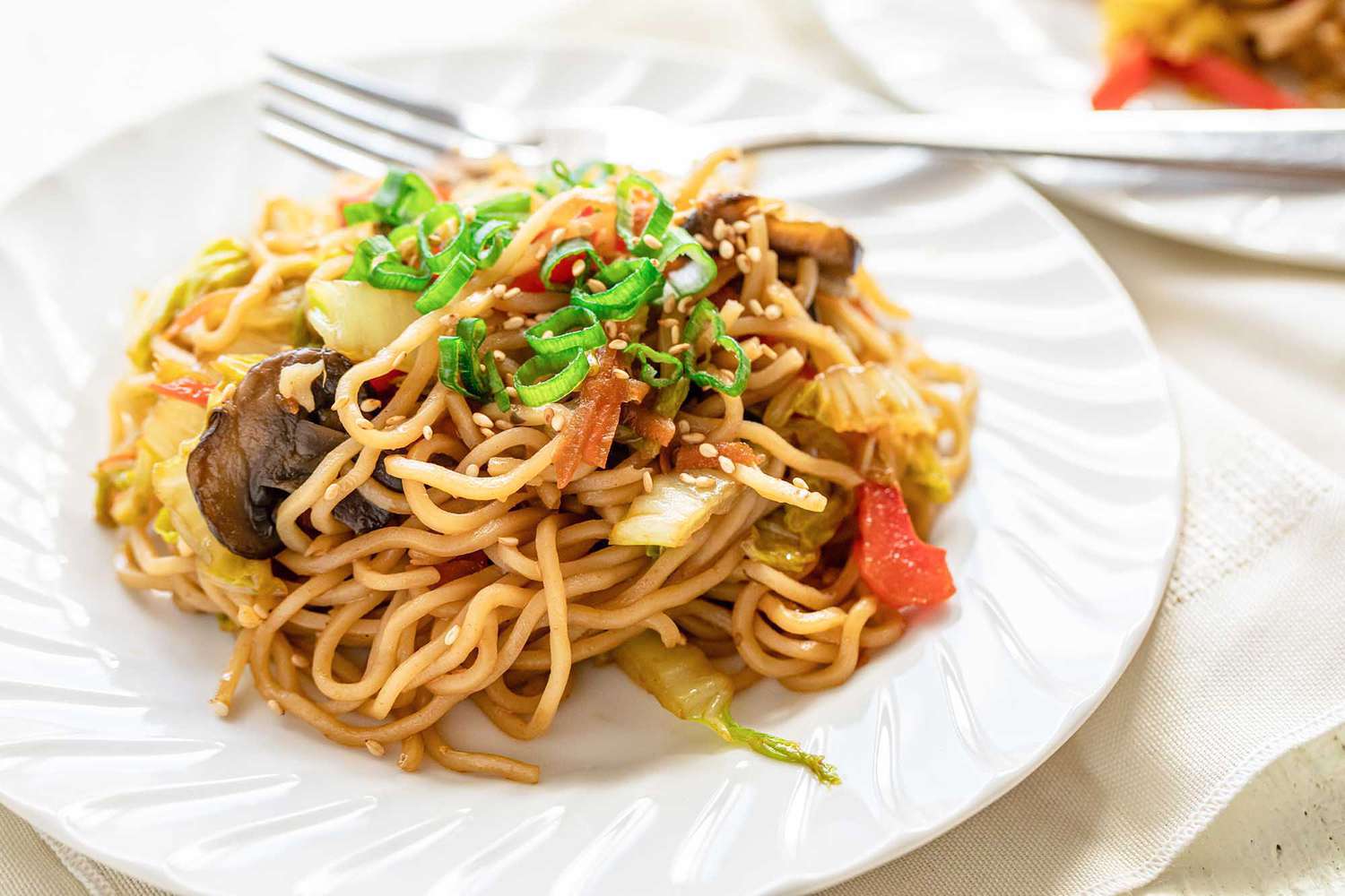 Side view of a white plate and fork with homemade vegetable lo mein. A fork is on the plate and a second plate is partially visible in the upper right hand corner.