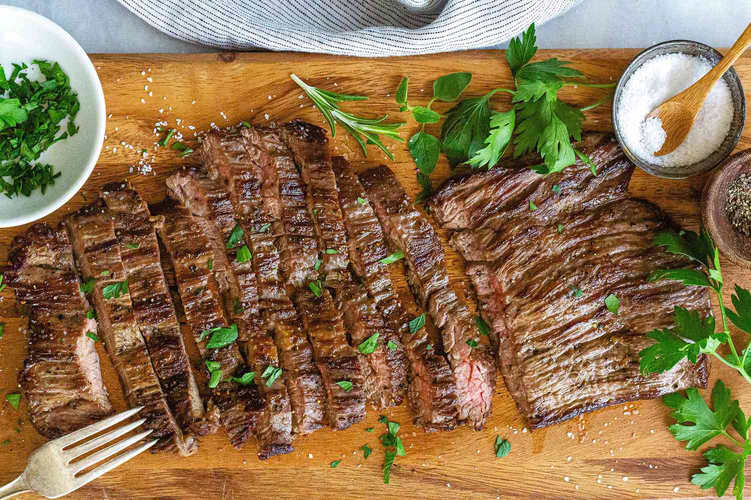 Sliced grilled skirt steak garnished with herbs served on a wooden cutting board accompanied by salt pepper and parsley