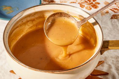 Angled view of a white pot of gravy sitting on a wooden trivet with a ladle with some gravy and all on a white and brown embroidered table cloth