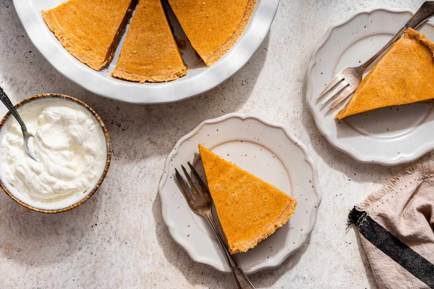 Slices of no-bake pumpkin pie on two small plates next to a bowl of whipped cream and a pie pan with more pumpkin pie