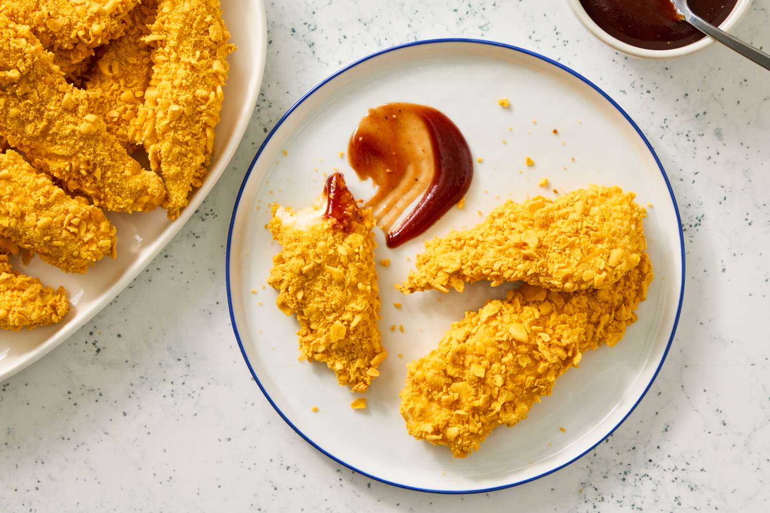 A closeup of breaded chicken tenders on a plate with some BBQ sauce