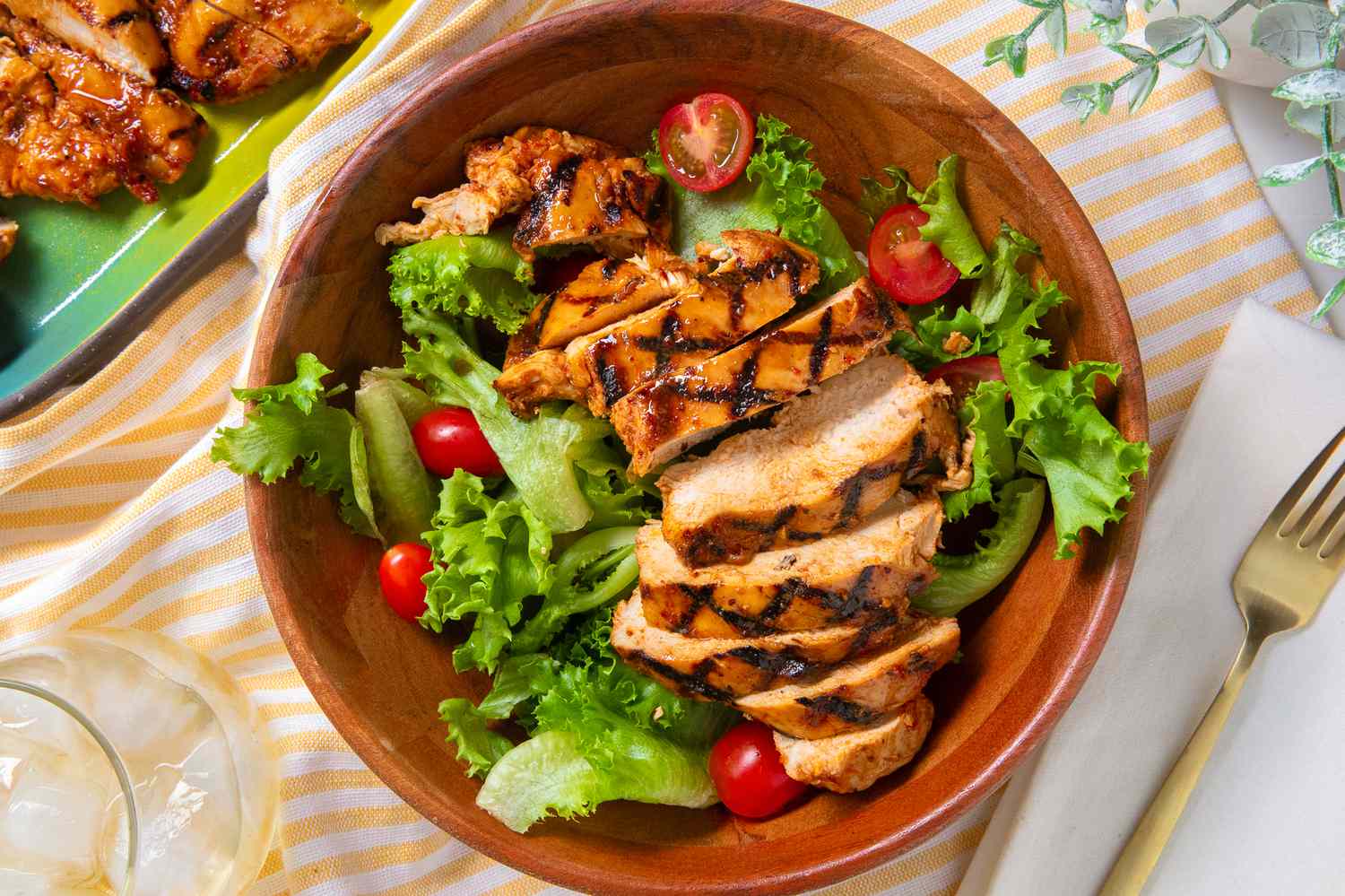 grilled chicken in a bowl over a salad at a table setting with more chicken on a platter, a glass of water, a fork, and a table plant