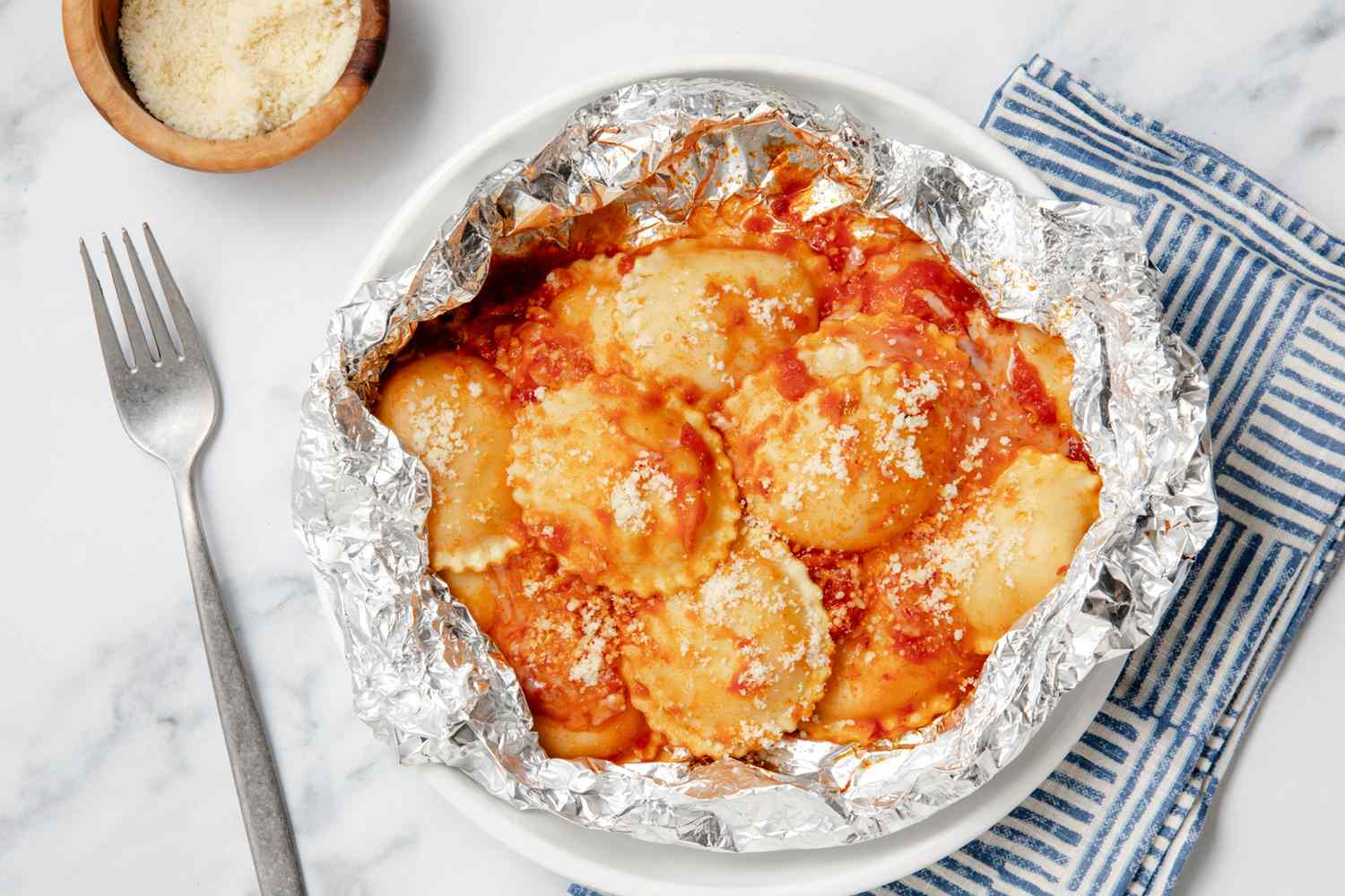 Foil-wrapped ravioli with tomato sauce and grated cheese, a fork and bowl of cheese nearby