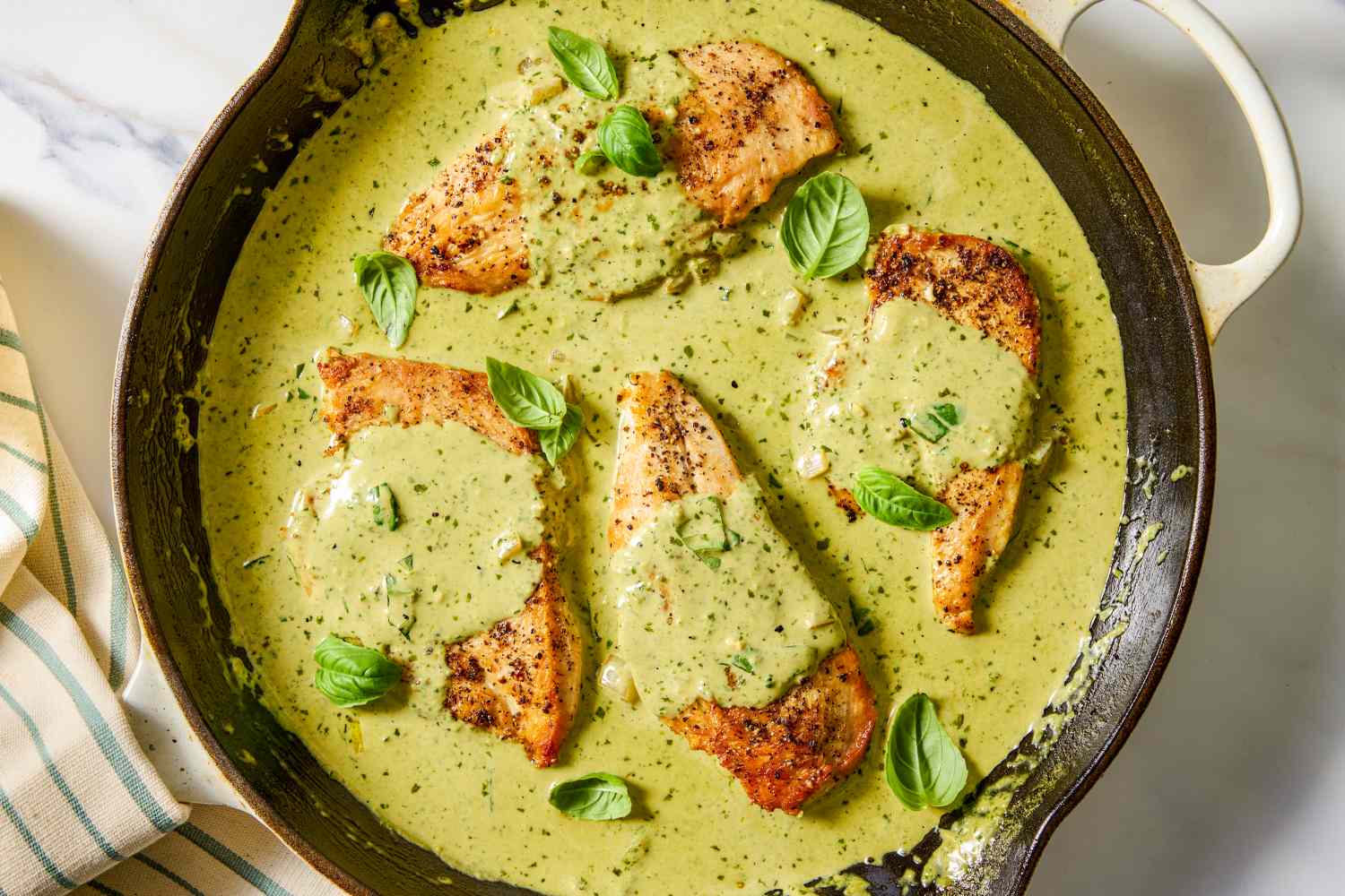 Four cooked chicken fillets in a skillet covered with green pesto sauce and garnished with basil leaves