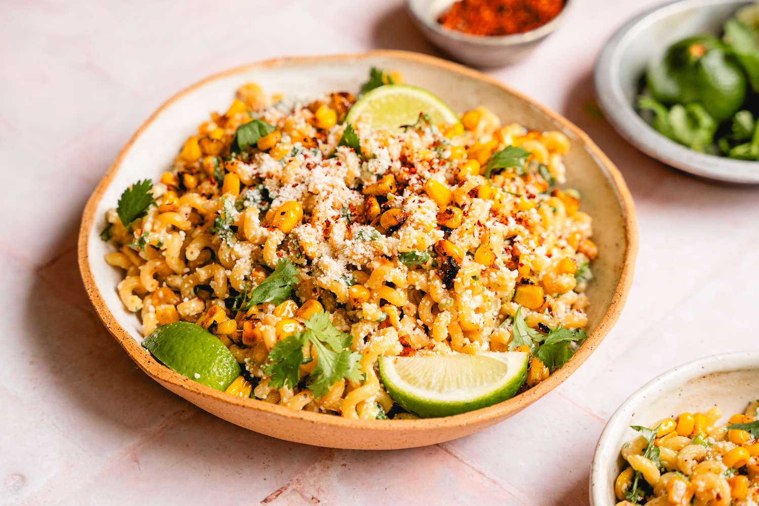A bowl of esquites pasta salad served with lime slices and garnished with herbs and cheese with seasoning bowls in the background