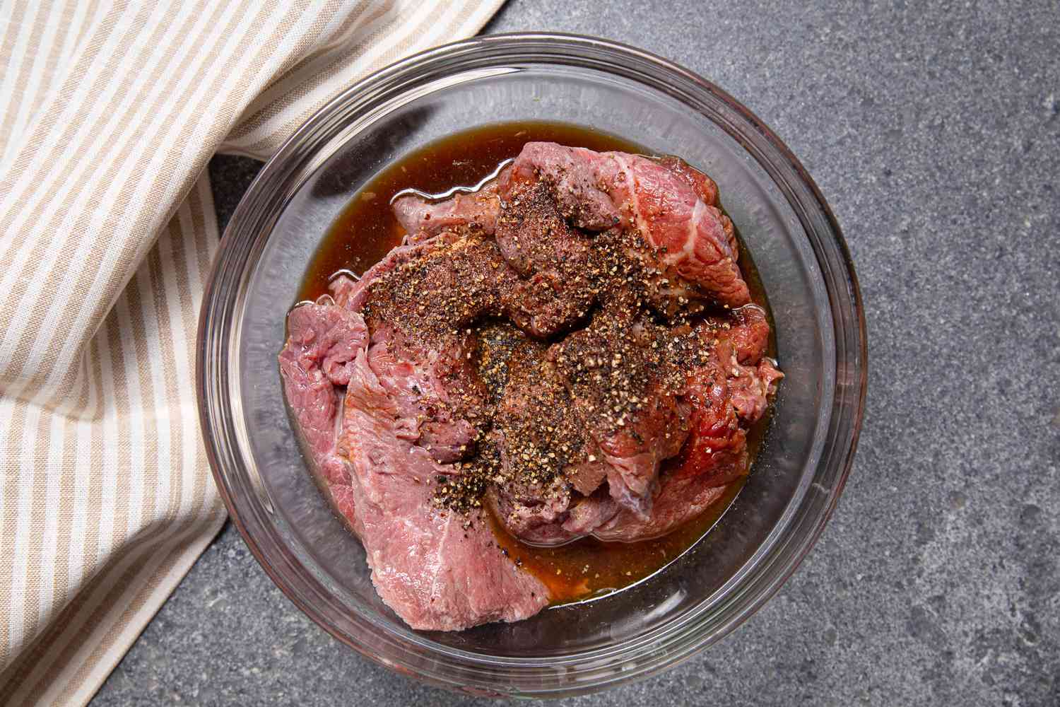 Beef Slices in a Bowl of Marinade Next to a Striped Kitchen Towel for Filipino Bistek Recipe