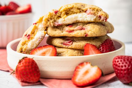 Stack of Strawberry Cheesecake Cookies With the Top Cookie Cut in Half to Showcase Cheesecake Filling in a Small Plate With a Few Halved Strawberries, and in the Surroundings, a Bowl of Strawberries and Some Free Flowing Strawberries (Some Whole, Some Halved) on the Counter, All on a Pink Table Napkin 