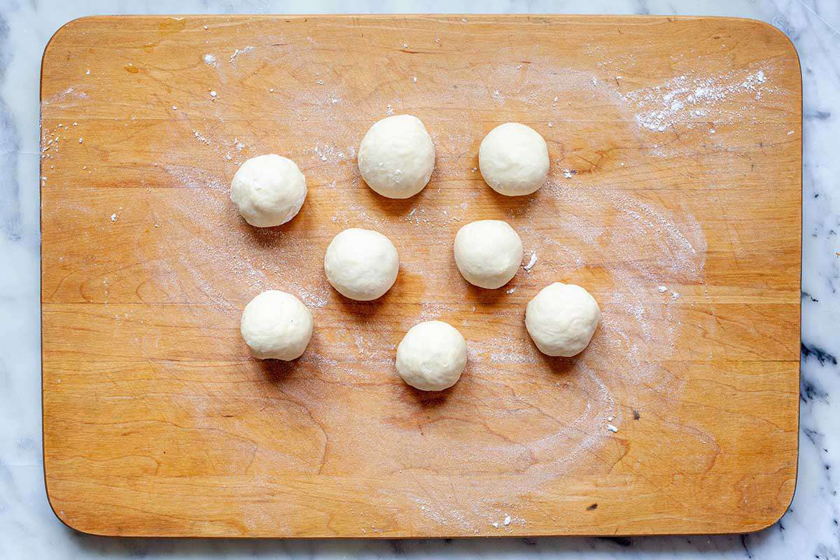 A wooden cutting board with eight dough balls for a flour tortilla recipe.