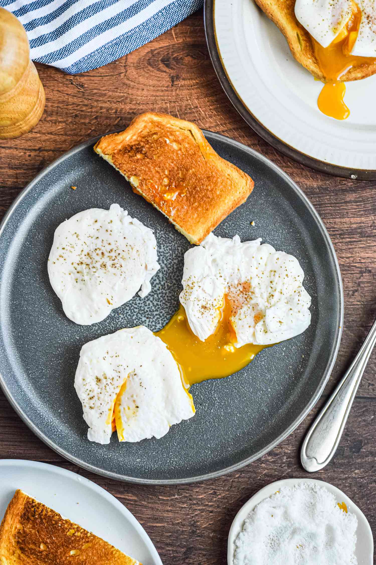 Three poached eggs and a piece of toast on a table with linens, plates, forks, and pepper grinder.