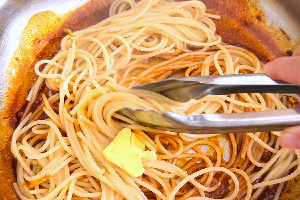 Pan with cooked noodles being stirred with tongs a pat of butter melting in the sauce