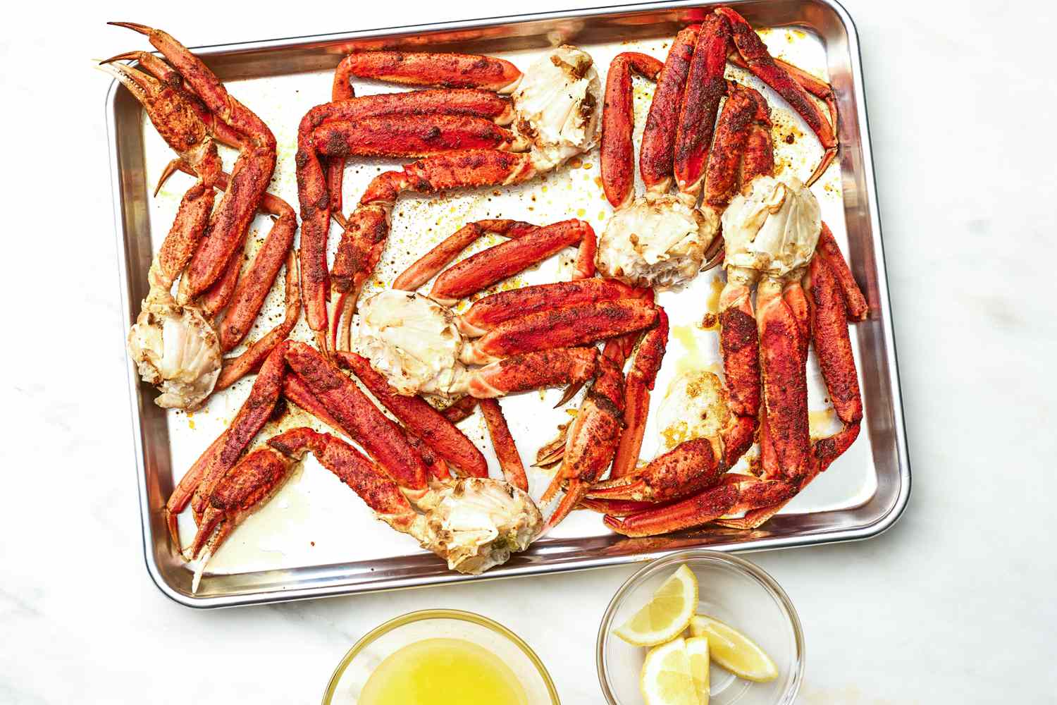 Baking Tray of Easy Baked Crab Legs, and on the Counter Next to It, a Bowl of Melted Butter and a Bowl of Lemon Wedges