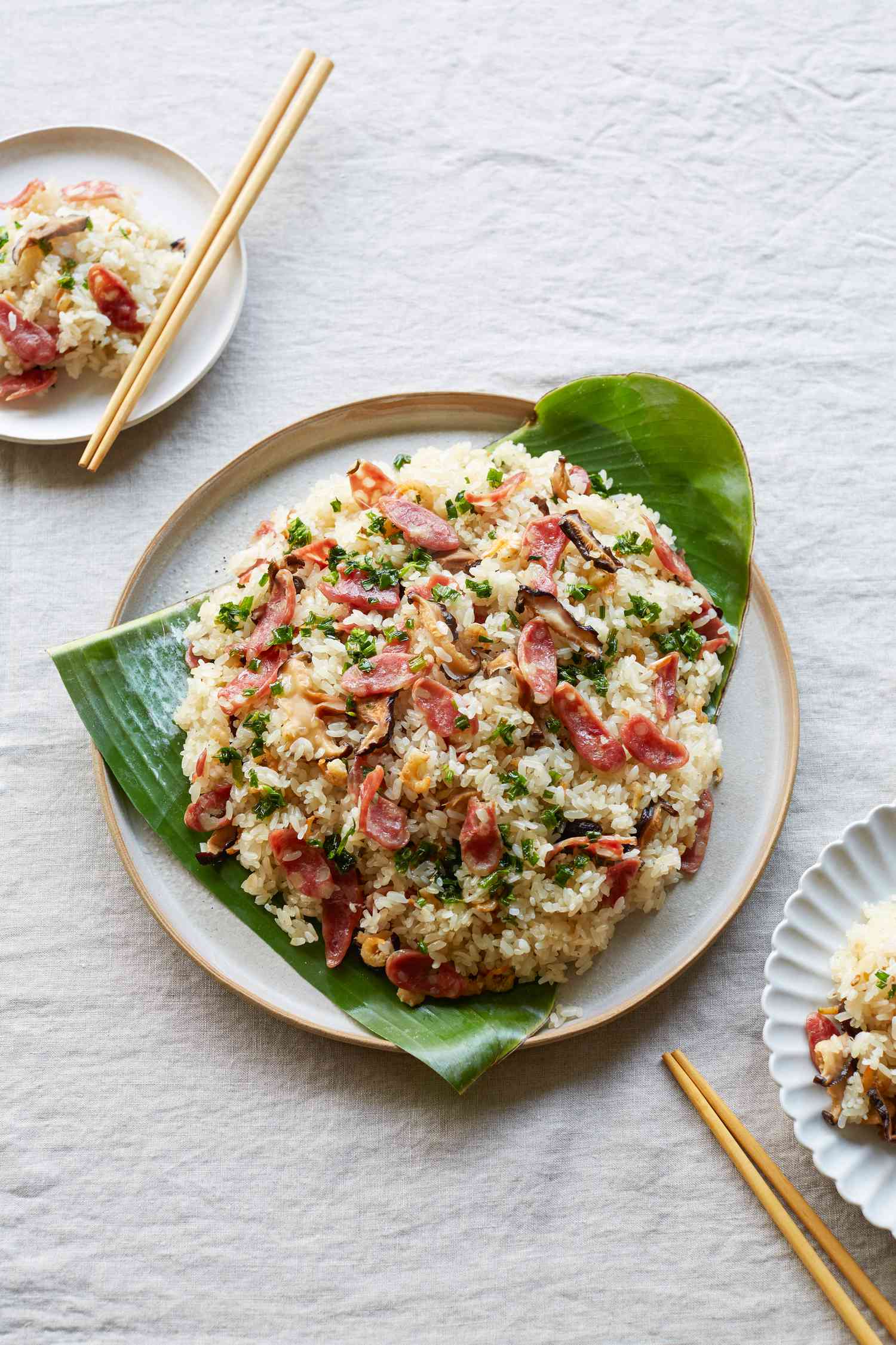 Sticky rice with lạp xưởng, shrimp, and mushrooms served over banana leaves and with chopsticks next to two smaller plates.