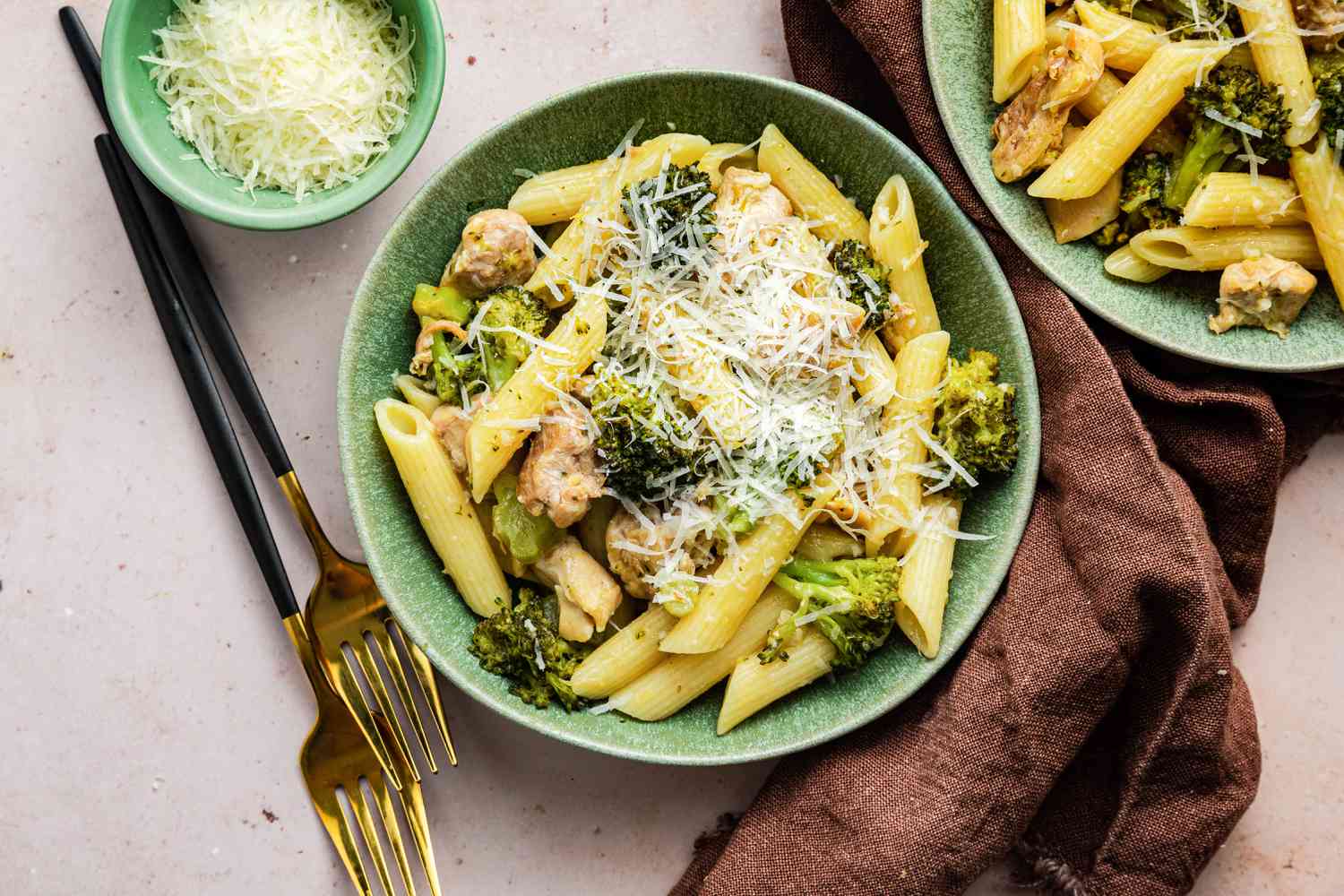 Two Bowls of Chicken and Broccoli Pasta Topped with Parmesan Cheese and Next to the Bowls, a Kitchen Towel, a Small Bowl of Cheese, and Some Utensils