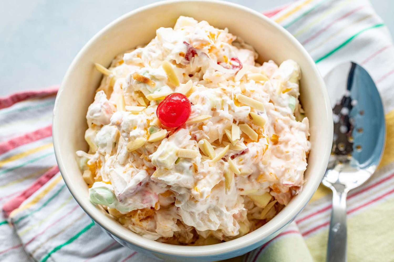 A mixing bowl of marshmallow salad is in the center of the horizontal image. Cream coated slivered almonds, mini marshmallows and fruit are in the bowl. A single bright red cherry is on the top of the salad. A striped dish towel is under the bowl and a slotted spoon is to the right.