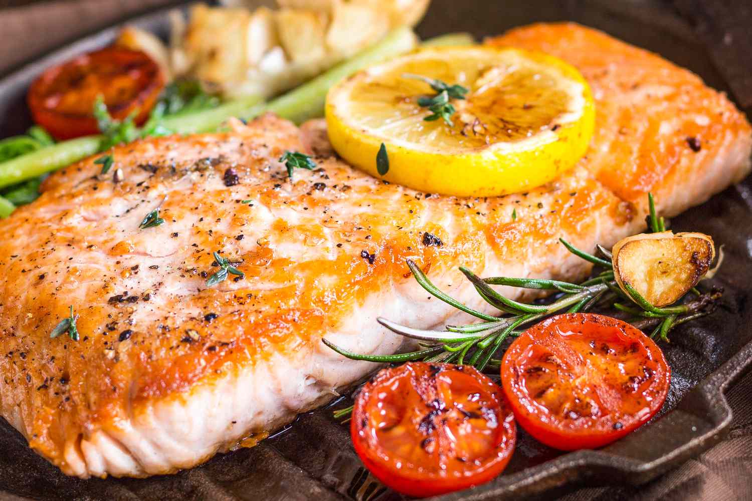 Close-up of a cooked salmon fillet in a grill pan with a lemon slice on top and a halved cherry tomato and rosemary sprig next to it