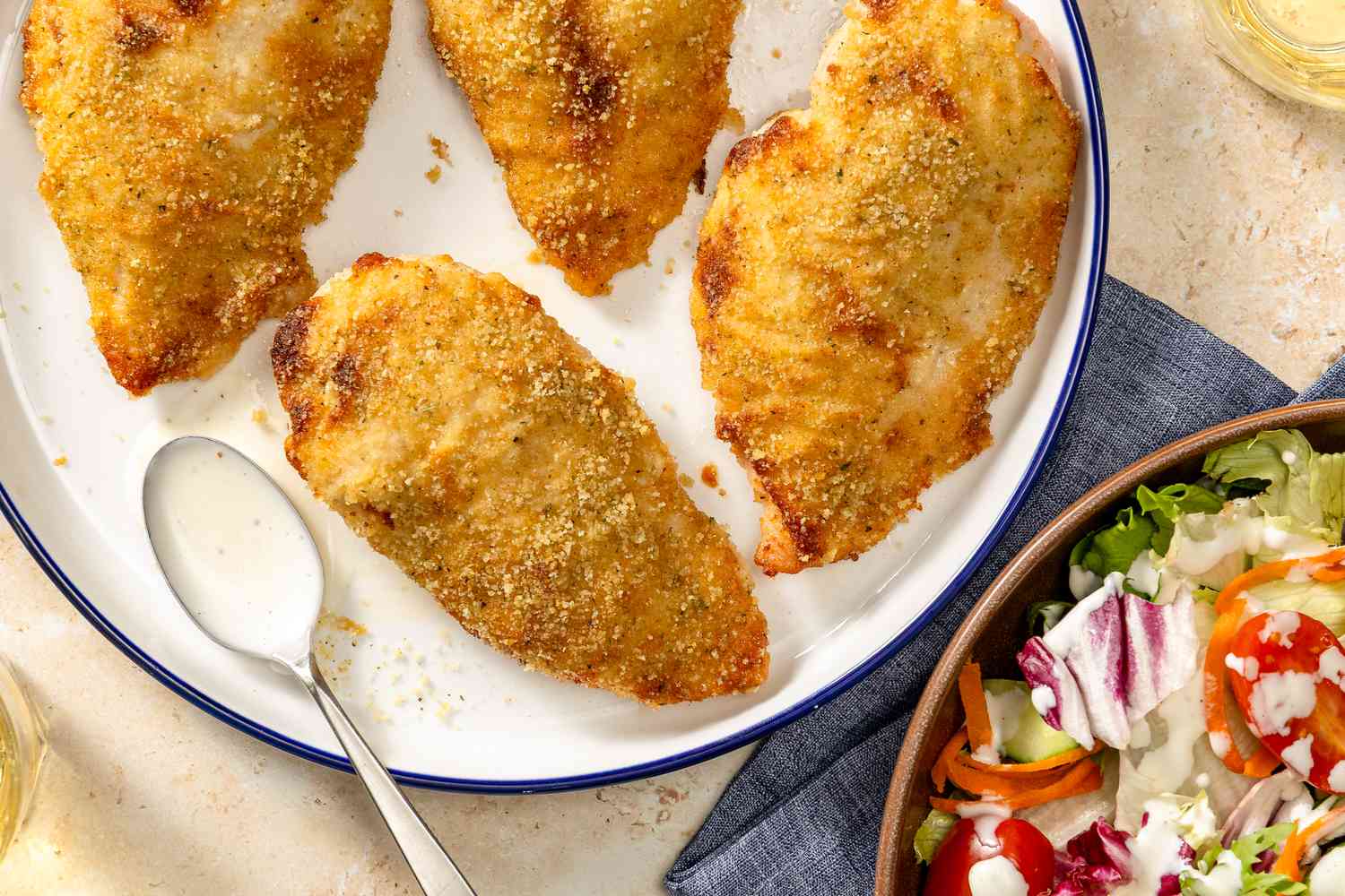Overhead view of a plate of ranch baked chicken breasts with a spoon of ranch dressing next to a bowl of salad