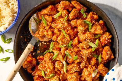General Tsos chicken dish with sesame seeds and green garnish in a skillet served with rice on the side in a bluerimmed bowl