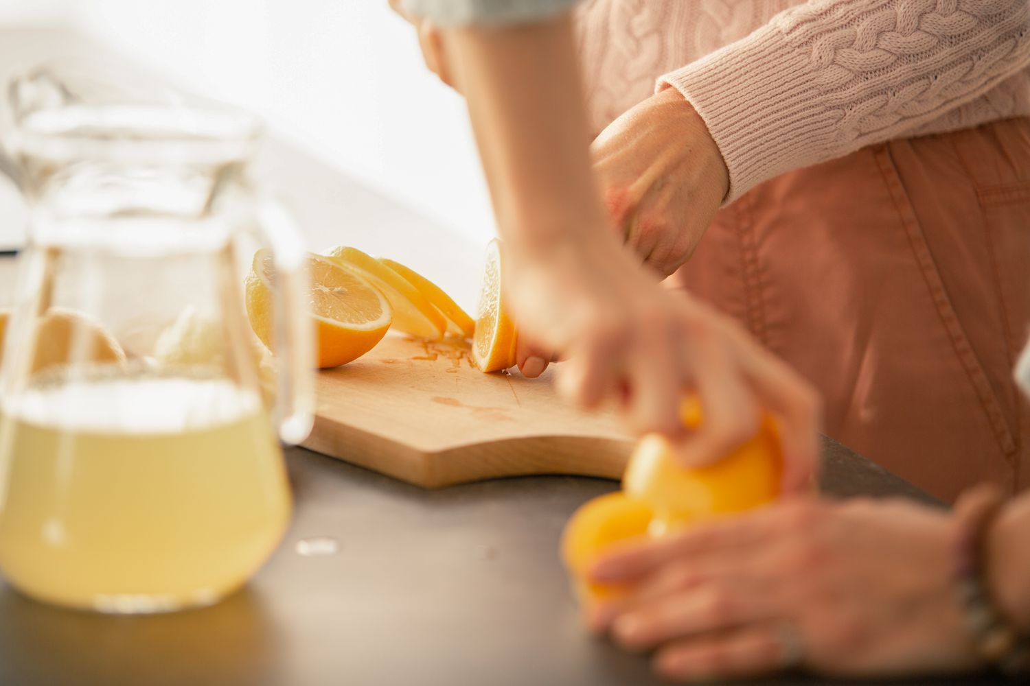 Hands cutting lemons on a wooden board with a pitcher of lemonade nearby