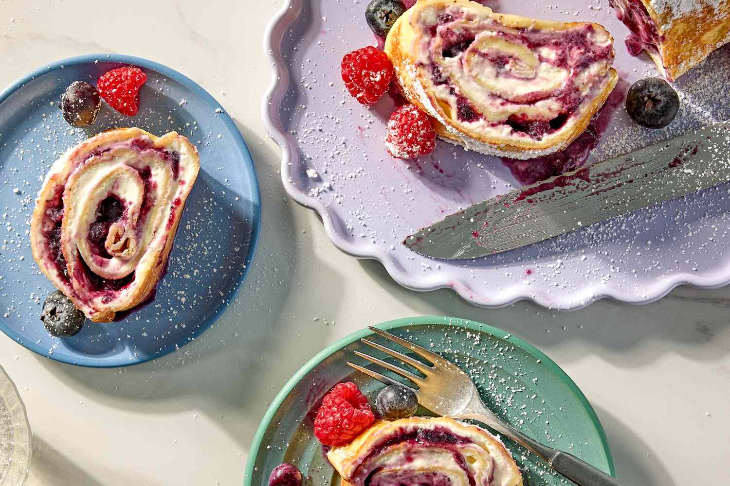 Slices of a rolled crepe cake filled with a fruit compote sprinkled with powdered sugar placed on colorful plates with small berries