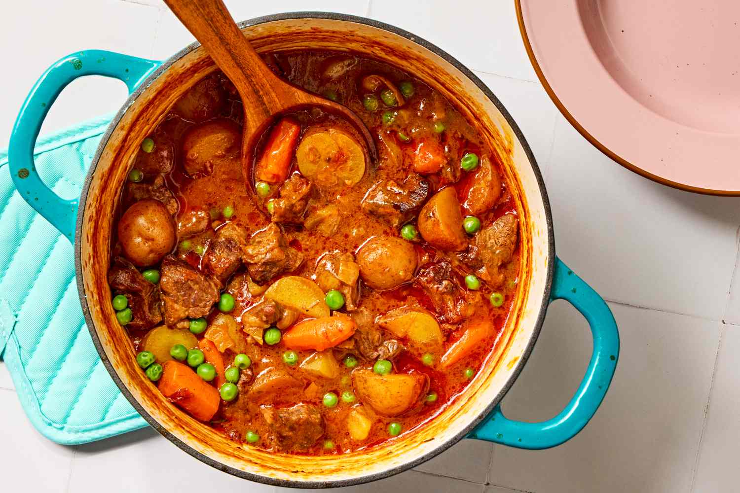 Overhead view of a teal colored dutch oven of beef stew and a wooden spoon all on a tile countertop next to a pink bowl and blue hotpad