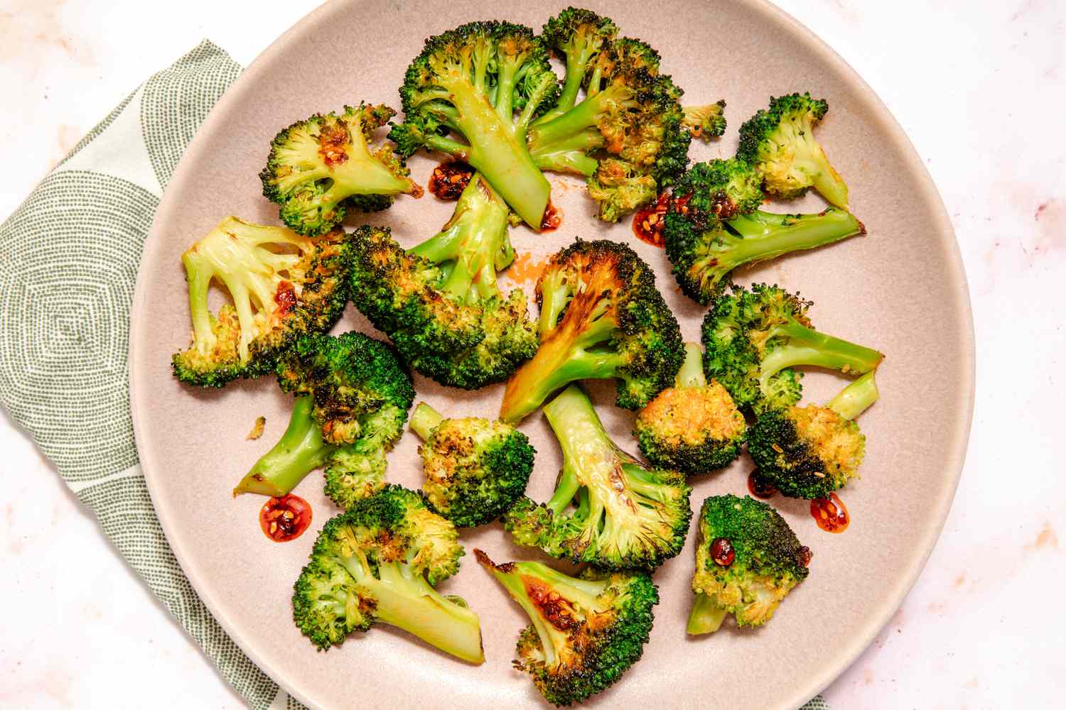 Overhead view of a pink plate of chili crisp coated broccoli florets resting on a cloth napkin and marble countertop