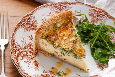A slice of best deep dish quiche on a decorative china plate. A handful of arugula is to the right of the quiche. A fork is to the left of the plate and a table linen is above the plate.
