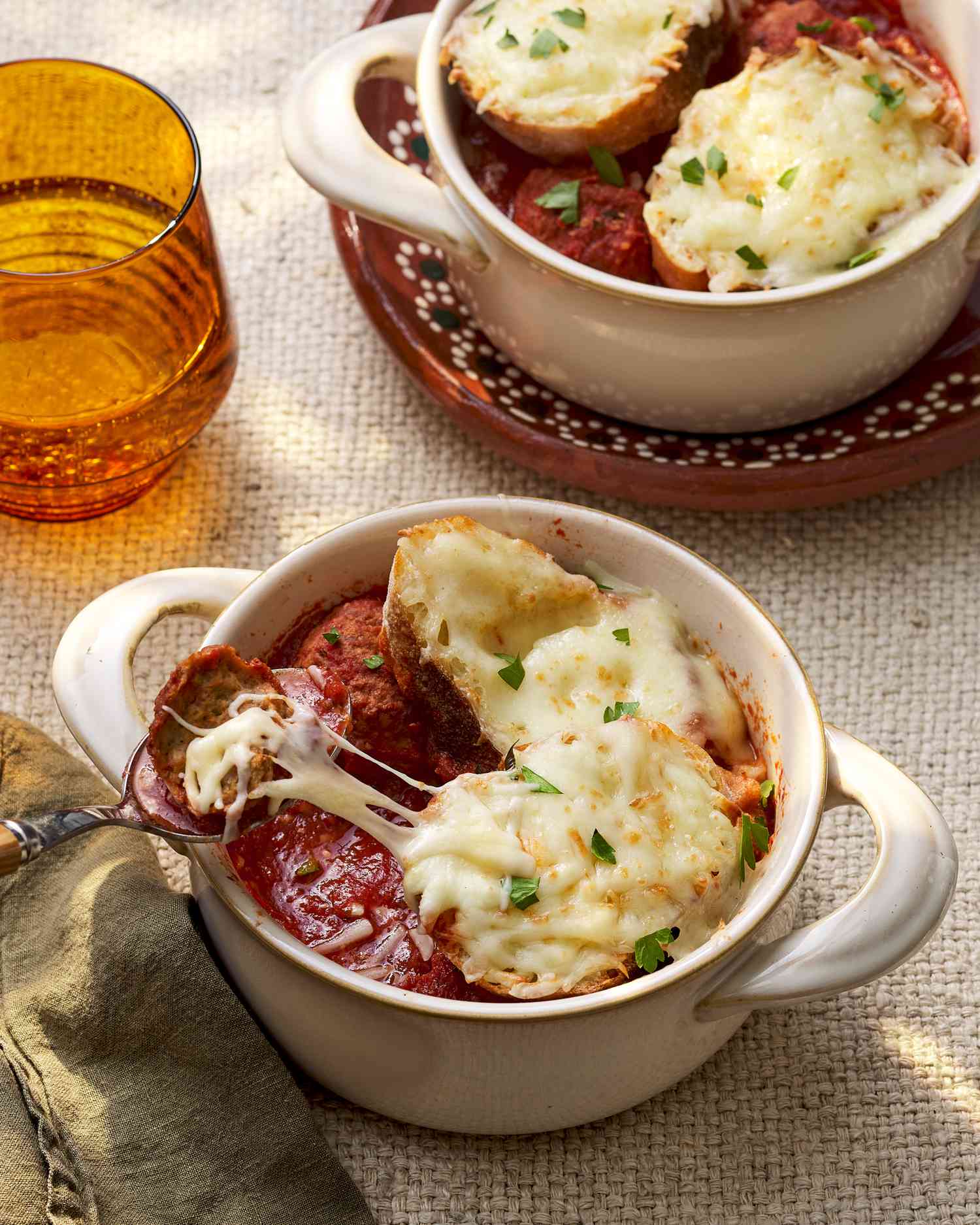 Meatball lifted from a pot-like ramekin, resulting in a cheese pull at a table setting with another pot-like ramekin, a glass of water, and a table napkin