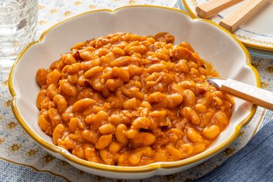 A bowl of baked beans with sauce and a spoon on a patterned tablecloth