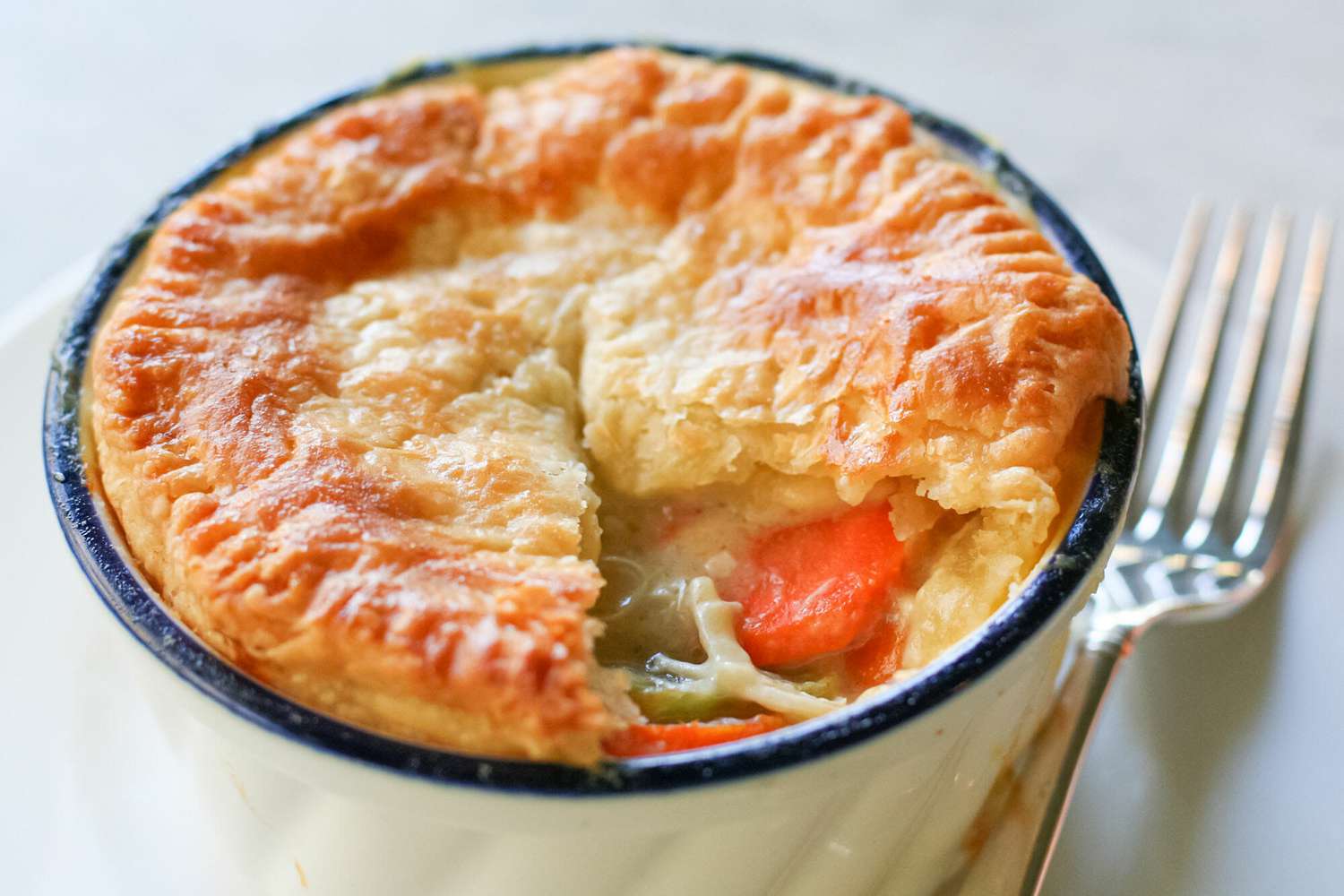 A pot pie with a golden crust is in a ramekin with some of the crust removed to show the filling.