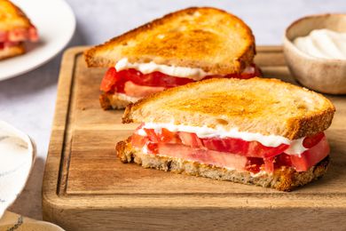 tomato sandwich halves on a cutting board next to a bowl of mayo and a plate with more tomato sandwiches