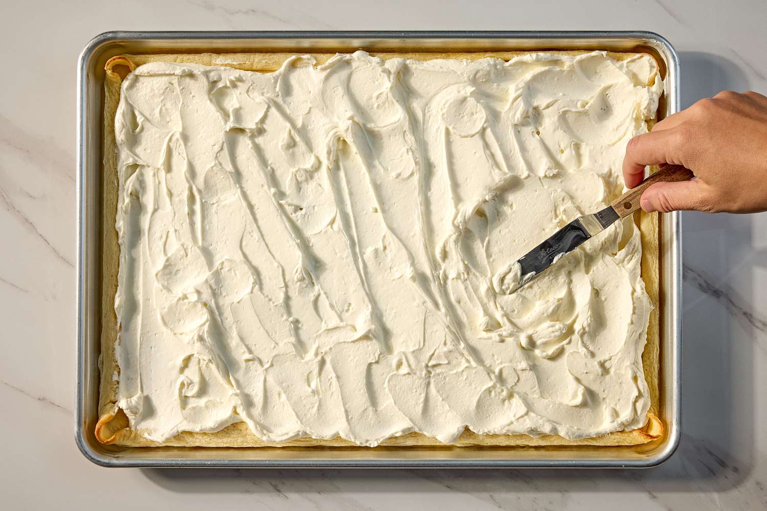 A hand spreading cream over a crepe cake layer in a rectangular pan with an offset spatula for the lazy crêpe cake recipe