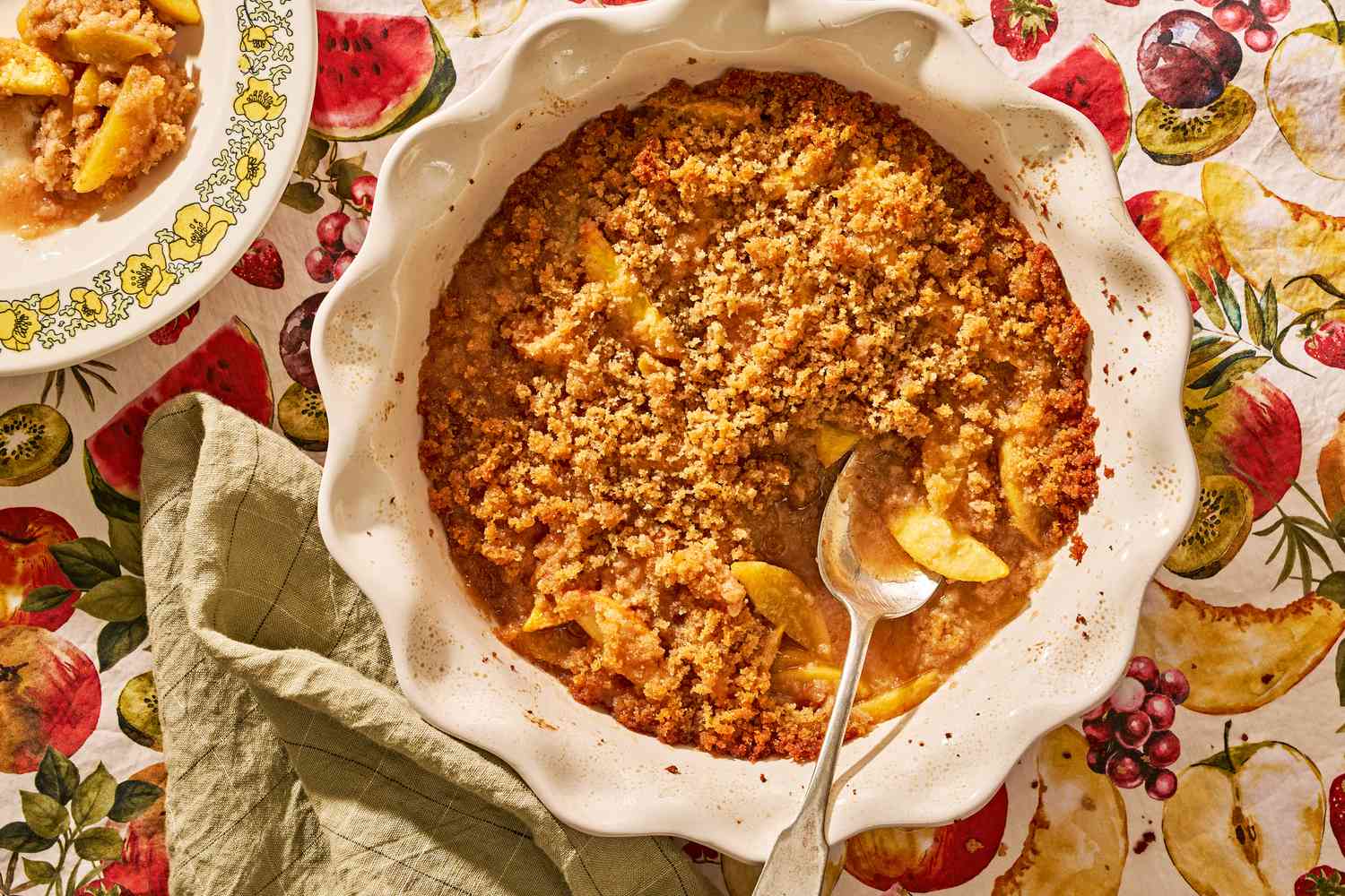 peach brown betty in a circular pie pan next to a serving in a bowl at a cozy table setting (tablecloth pattern is watercolor cut fruit)