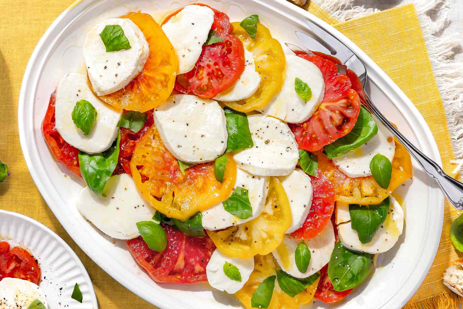 Platter of Caprese Salad With Tomatoes, Basil, and Mozzarella Next to a Plate With a Serving, All on a Yellow Placemat
