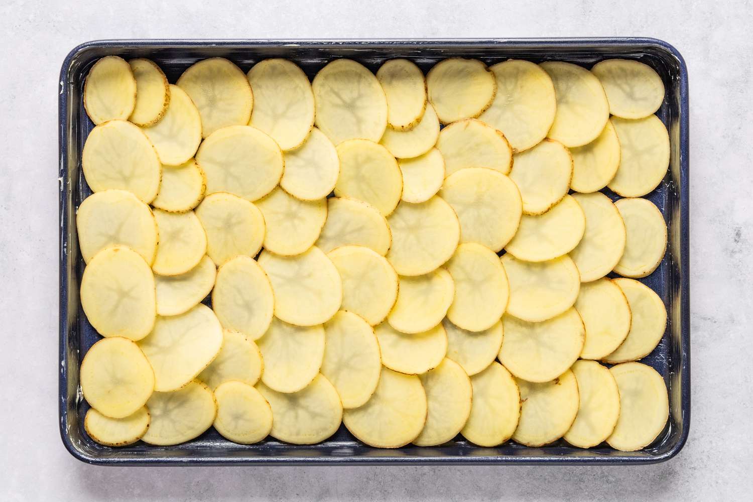 Overhead view of a 9x13 sheet pan with slices of potato shingled across the sheet for Sheet Pan Au Gratin Potatoes recipe