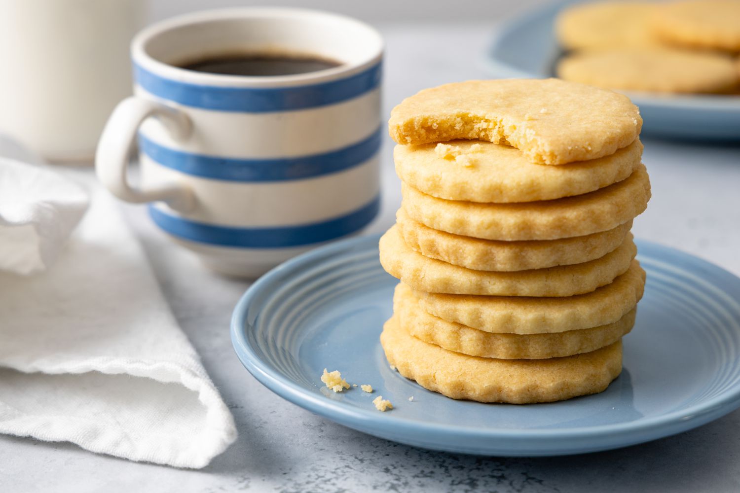 Stack of Classic Shortbread Cookies with the Top Cookie Missing a Bite and a Cup of Coffee in the Background